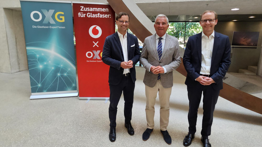 Drei Männer in formeller Kleidung stehen nebeneinander vor zwei Rollups mit den Logos von OXG und Vodafone in einer modernen Innenarchitektur.