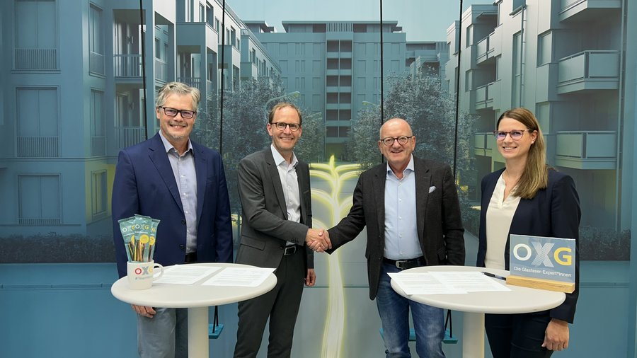 Personen in professioneller Kleidung schütteln sich die Hände vor einem modernen Hintergrund mit Wohngebäuden, während sie an runden Tischen stehen, die mit Dokumenten und Unternehmenssymbolen dekoriert sind