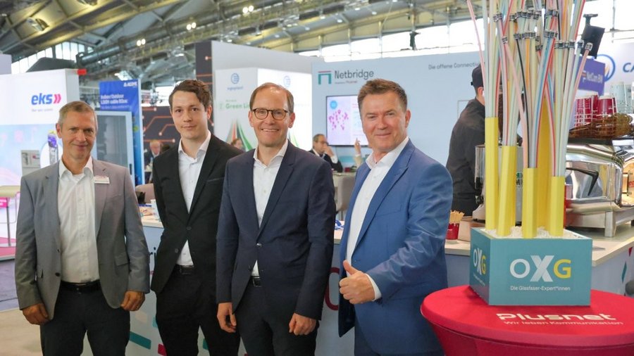Vier Personen stehen nebeneinander an einem Messestand in einer Ausstellungshalle. Im Vordergrund ist ein roter Stehtisch mit Plusnet‑Logo sowie eine Kabelinstallation mit OXG‑Logo zu sehen, im Hintergrund weitere Messestände und Besucher.