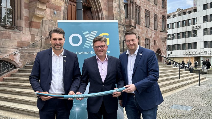 Drei Männer stehen vor einem historischen Gebäude in Saarbrücken und halten gemeinsam ein Stück Glasfaserkabel, während im Hintergrund ein Banner der Firma OXG zu sehen ist.
