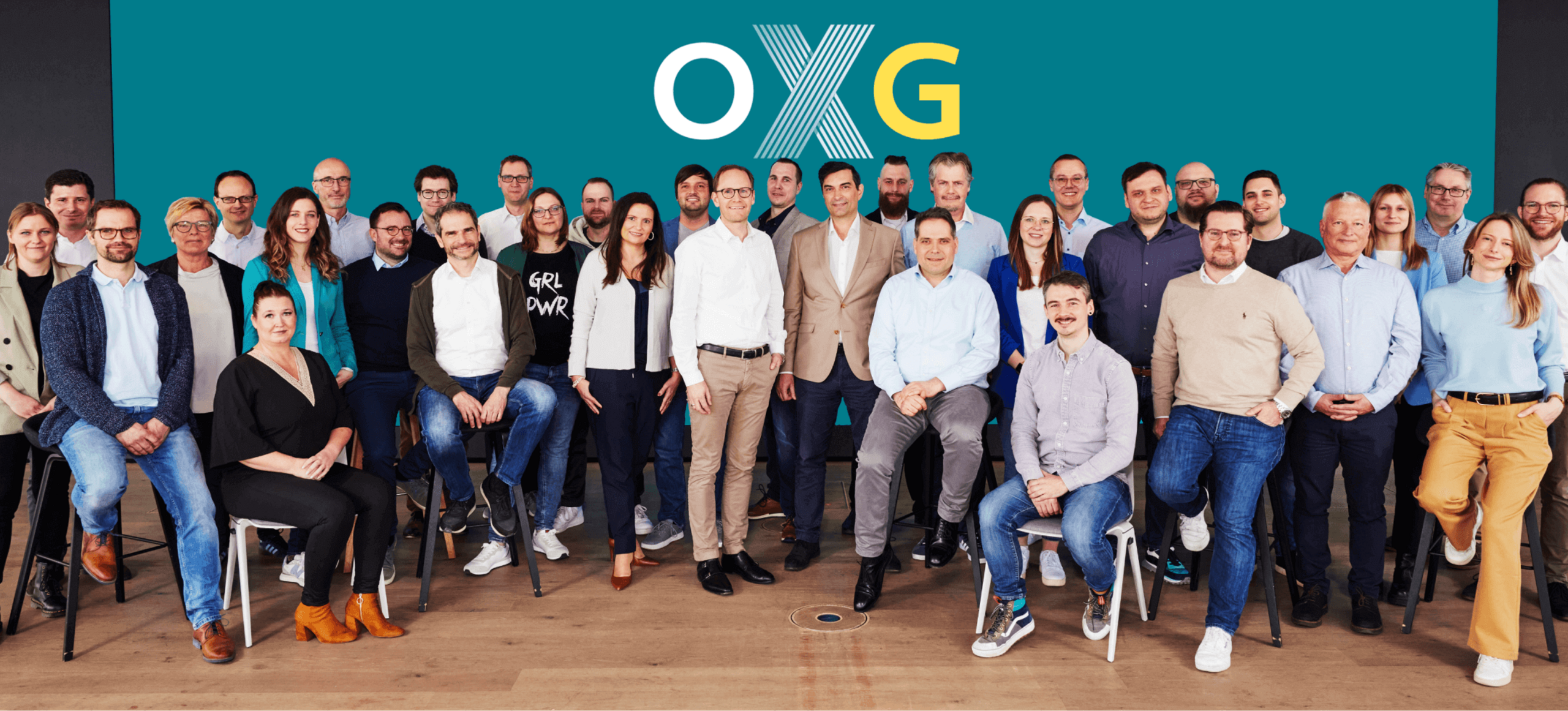 Gruppenfoto des OXG Glasfaser Teams im Büro, bestehend aus 32 Personen in formeller und legerer Kleidung vor einer türkisfarbenen Wand mit dem OXG-Logo