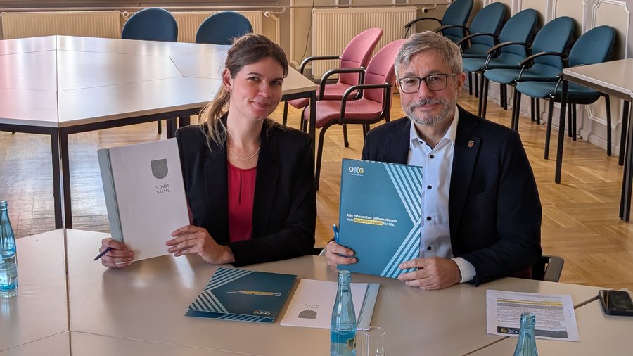 Zwei Personen sitzen an einem Tisch und halten Dokumente zum Glasfaserausbau in Suhl in den Händen, während im Hintergrund Stühle und ein größerer Tisch sichtbar sind