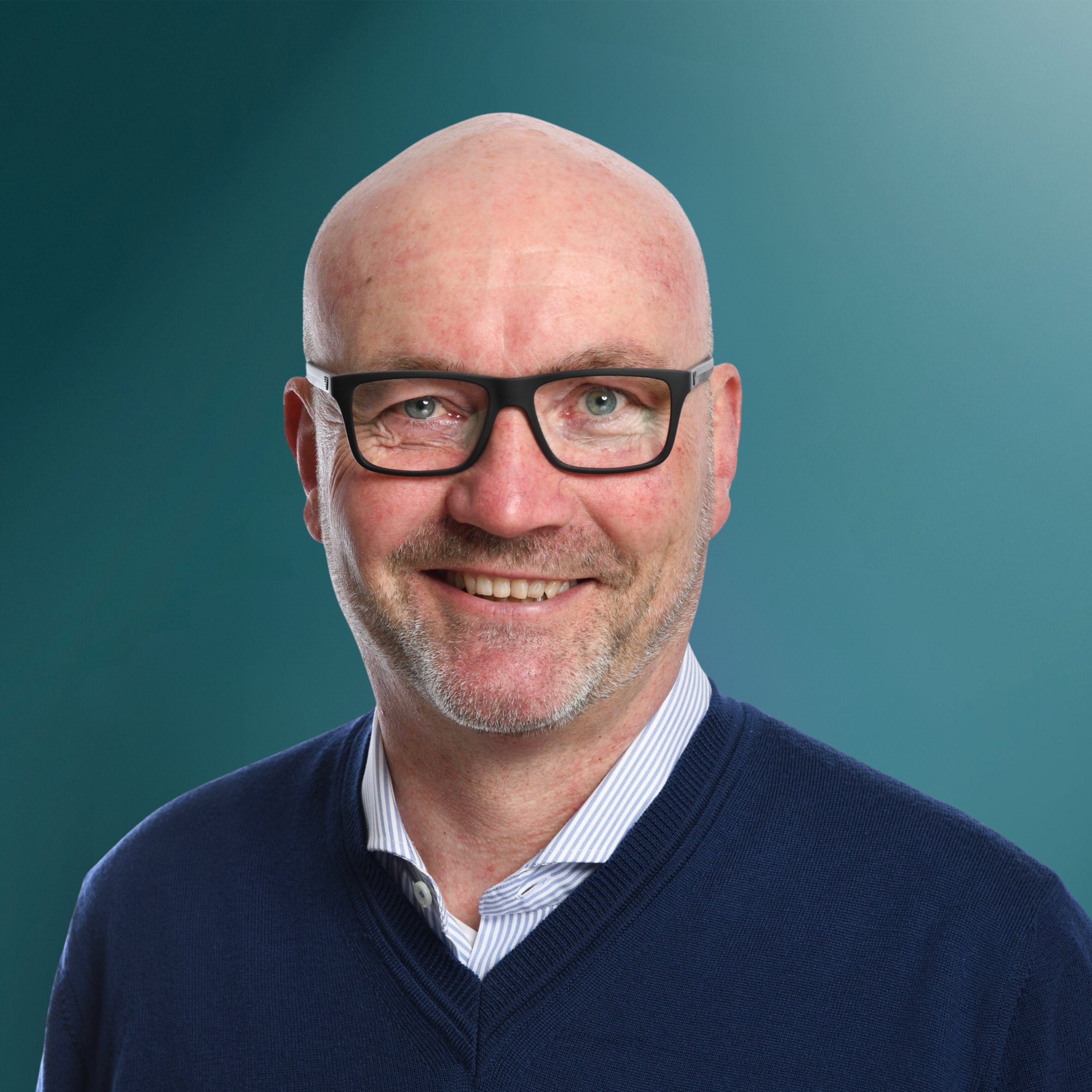 Portrait eines lächelnden Mannes mit Glatze und Brille, der einen blauen Pullover und ein gestreiftes Hemd trägt, vor einem blauen Hintergrund