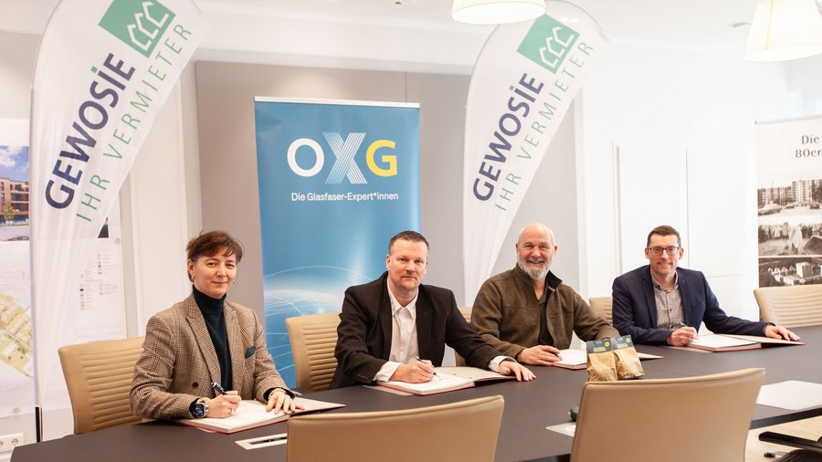 Besprechungsraum mit vier Personen, die an einem Tisch sitzen und Verträge unterzeichnen, umgeben von Werbebannern von GEWOSIE und OXG