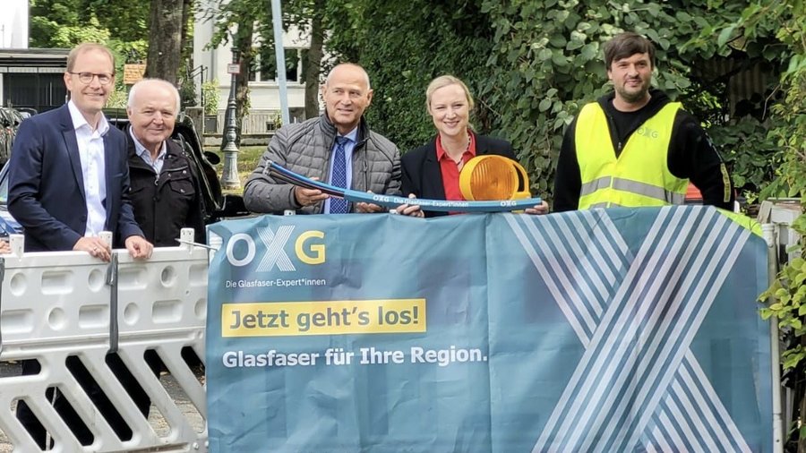 Fünf Personen stehen vor einem Zaun in der Stadt Mainz und halten ein Glasfaserkabel sowie ein Warnlicht, während sie vor einem Banner mit der Aufschrift "Jetzt geht's los! Glasfaser für Ihre Region" posieren.