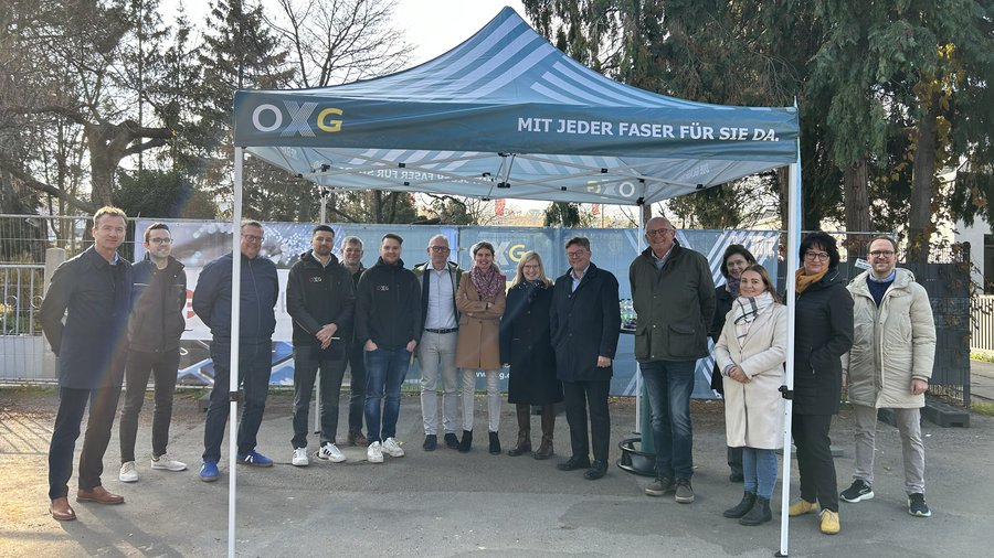 Eine Gruppe von Personen steht unter einem Banner der Marke OXG, das den Slogan „Mit jeder Faser für Sie da“ trägt, während sie in einem Außenbereich posieren.