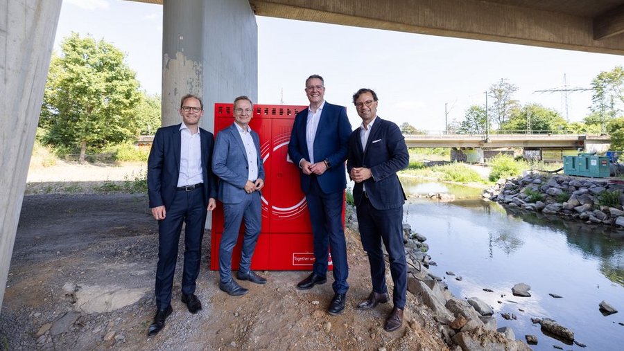 Vier Männer in Anzügen stehen unter einer Brücke neben einem roten Schrank, während im Hintergrund ein Gewässer und eine grüne Landschaft sichtbar sind.