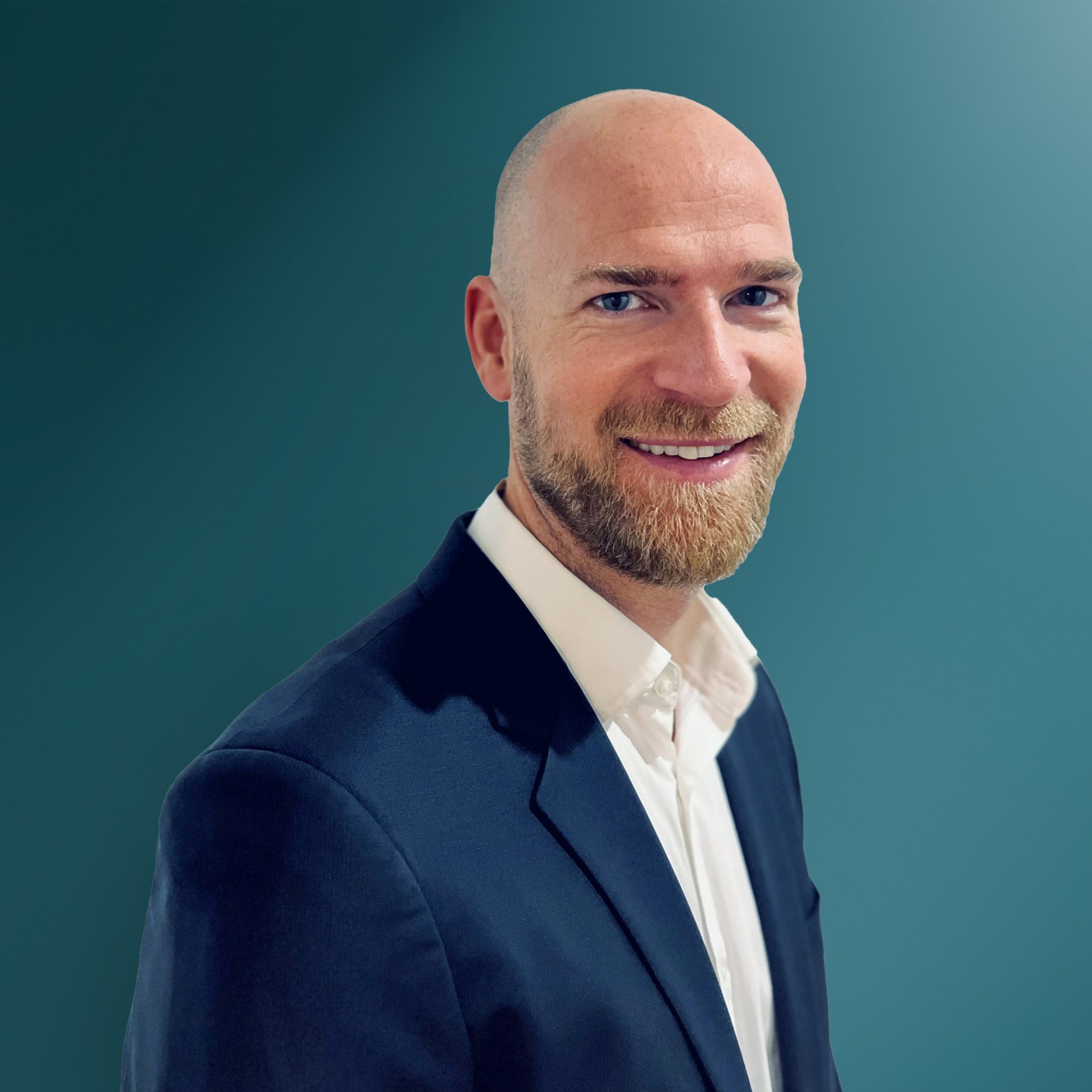 Ein lächelnder Mann mit kurzer, blonder Haar und Bart trägt einen schwarzen Anzug und ein weißes Hemd vor einem grün-blauen Hintergrund