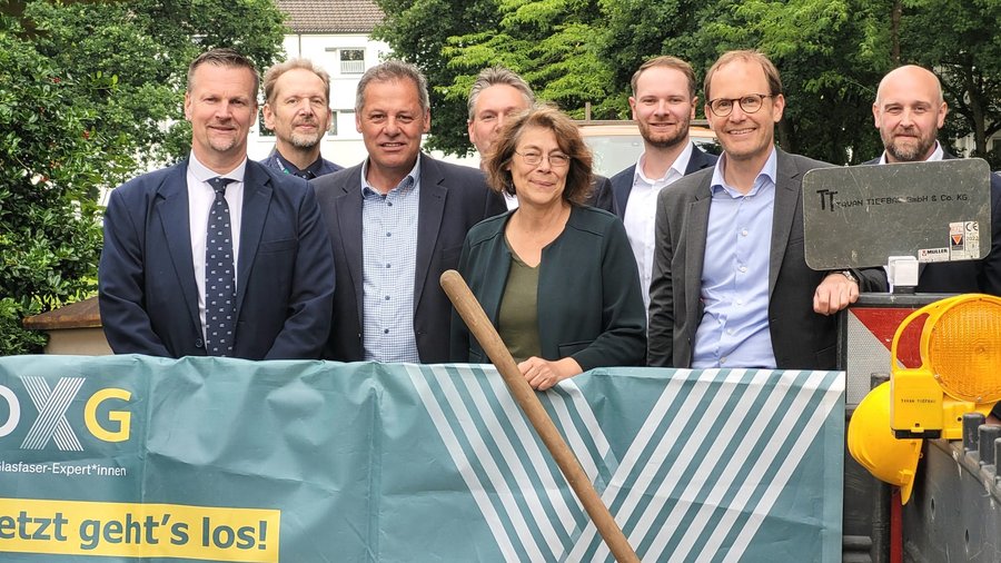 Gruppenfoto von acht Personen in Bremen, die vor einem Bauzaun mit einem Banner stehen, auf dem "Jetzt geht's los!" steht, während eine Frau einen Spaten hält