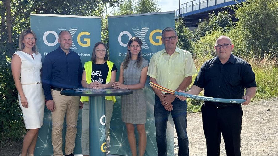 Gruppenfoto von sechs Personen, die vor zwei Banner mit dem Logo von OXG stehen und eine Glasfaserleitung halten, umgeben von einer naturnahen Umgebung in Torgau