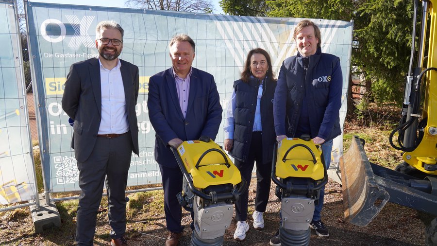 Vier Personen stehen draußen auf einer Baustelle vor einem Bauzaun mit OXG‑Banner, zwei von ihnen halten gelbe Verdichtungsgeräte, daneben steht ein Bagger.