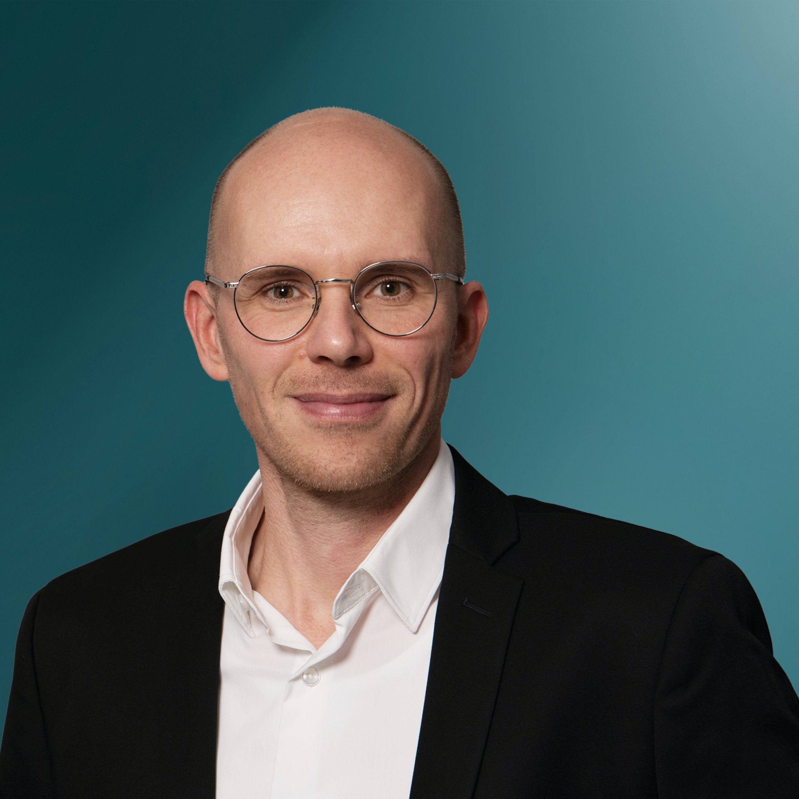 Porträt eines lächelnden Mannes mit Gläsern, der einen schwarzen Anzug über einem weißen Hemd trägt, vor einem einfarbigen, sanft strukturierten Hintergrund in Türkis- und Blautönen