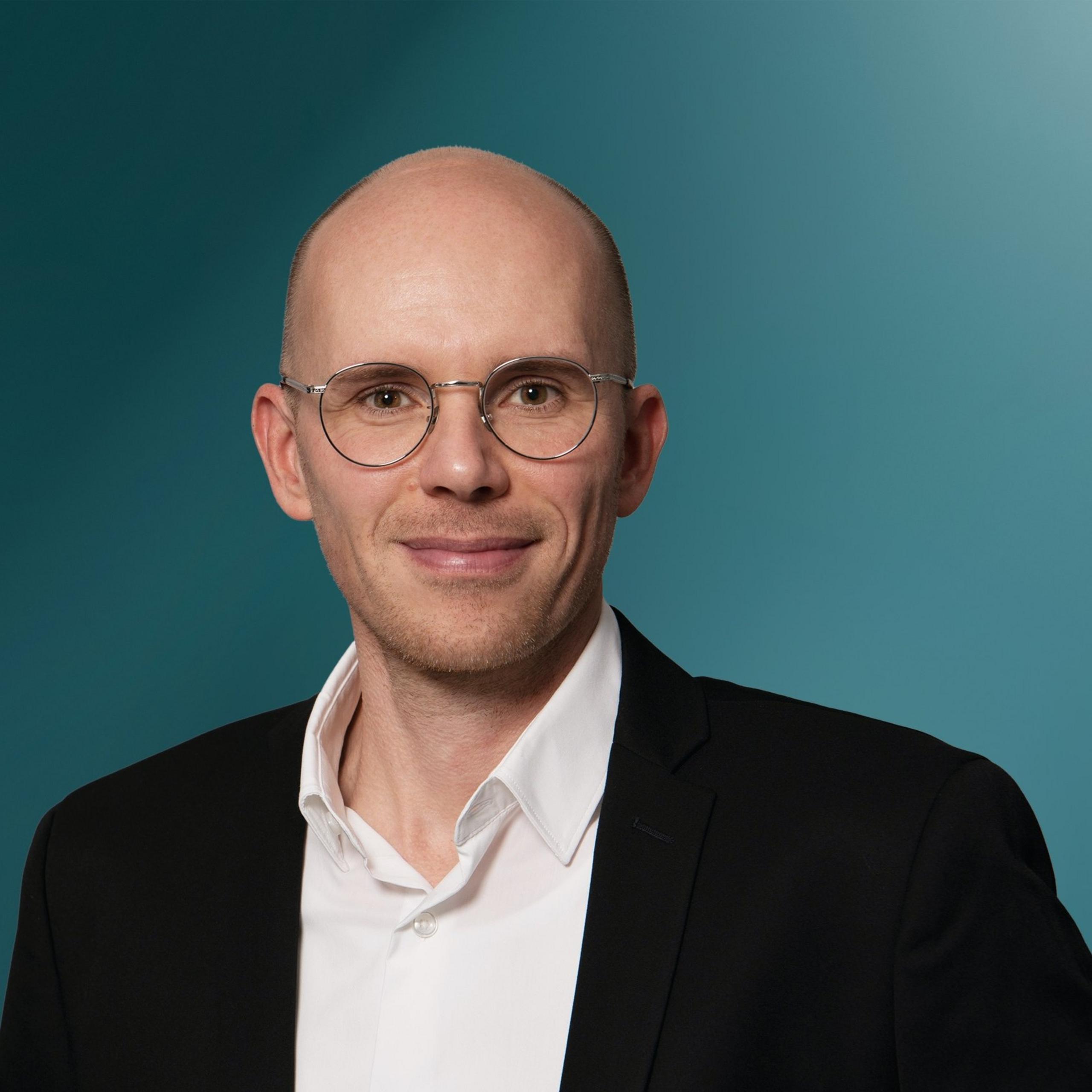 Porträt eines lächelnden Mannes mit Gläsern, der einen schwarzen Anzug über einem weißen Hemd trägt, vor einem einfarbigen, sanft strukturierten Hintergrund in Türkis- und Blautönen