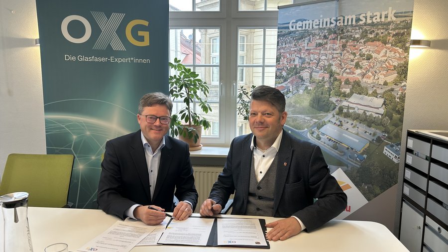 Zwei Männer sitzen an einem Tisch und unterzeichnen Dokumente zum Glasfaserausbau in Zittau, während im Hintergrund ein Banner mit dem Aufdruck „Gemeinsam stark“ und das Logo der Firma OXG sichtbar sind.