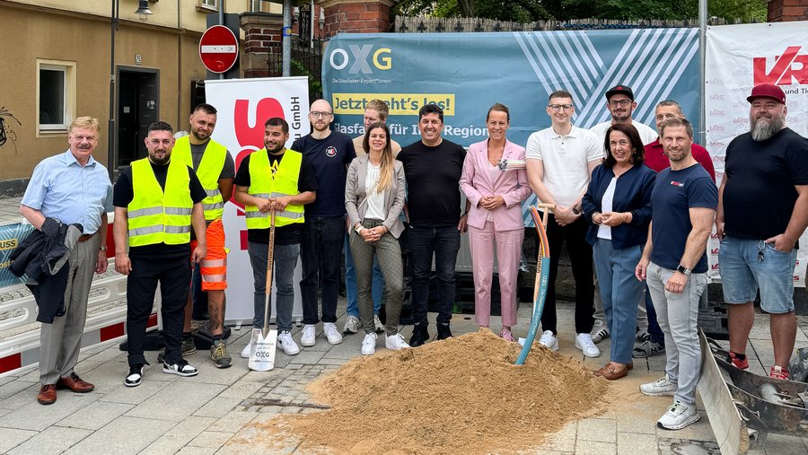 Gruppenporträt von Mitarbeitern und Gästen bei der Grundsteinlegung eines Glasfaserprojekts in der Stadt Hof, umgeben von Baumaterialien und Werbebannern