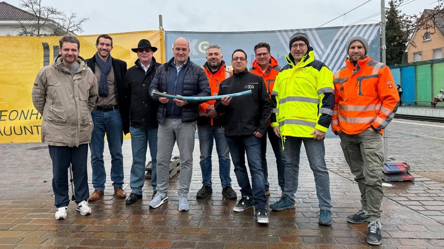 Gruppe von Personen in Arbeitskleidung, die ein farbiges Rohr halten, vor einem Banner mit der Aufschrift "OXG" und einer städtischen Kulisse der Stadt Dossenheim im Hintergrund