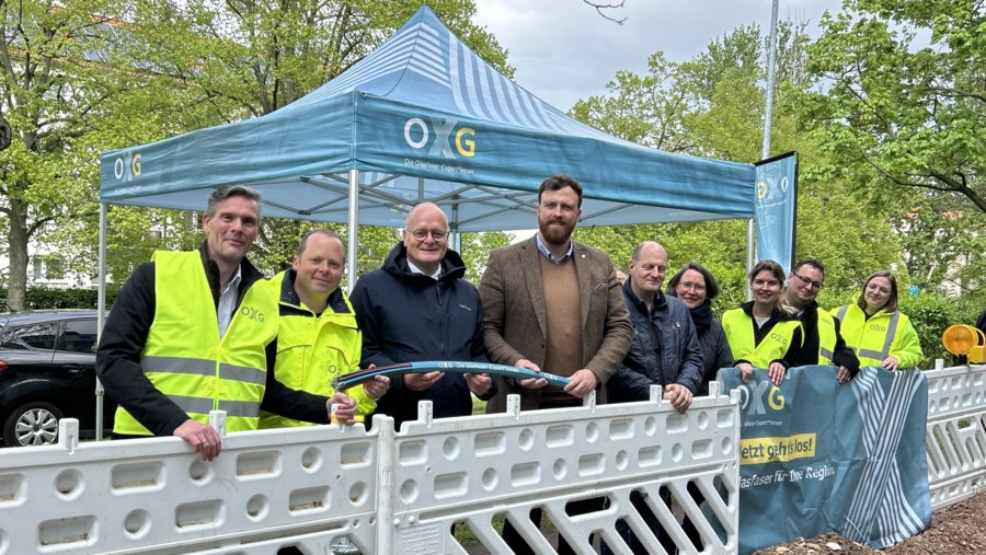 Gruppenfoto von Mitarbeitern der OXG, die im Freien stehen und eine Glasfaserleitung präsentieren, während sie in leuchtenden Sicherheitswesten gekleidet sind und hinter einer Absperrung stehen