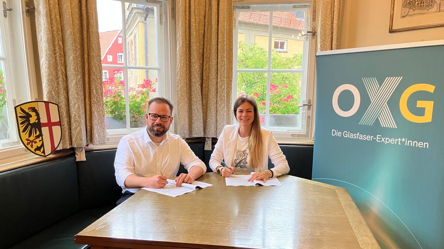Zwei Personen signieren Verträge an einem Tisch, während im Hintergrund ein Roll-up-Banner der Firma OXG und das Wappen der Stadt Memmingen zu sehen sind.