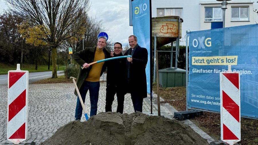 Drei Personen posieren in der Stadt Ravensburg mit einem Schaufel und einem Kabel vor einem Baustellenbanner, während im Hintergrund eine Erdeinehmtstelle sichtbar ist.