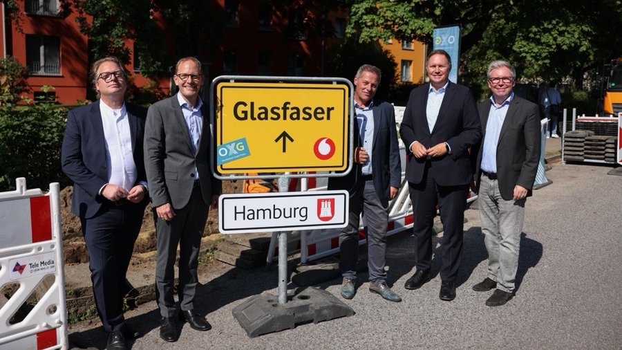 Fünf Männer in Anzügen stehen zusammen vor einem Verkehrsschild mit der Aufschrift "Glasfaser" in Hamburg, umgeben von Baustellenatmosphäre.