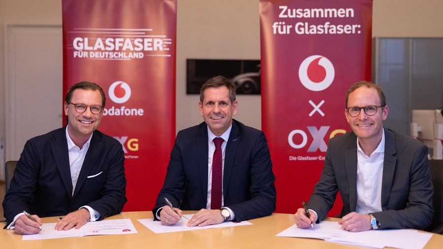 Drei Männer sitzen an einem Tisch und unterzeichnen Verträge, während im Hintergrund Banner von Vodafone und OXG für Glasfaser stehen