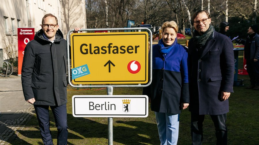 Drei Personen posieren vor einem Verkehrsschild mit der Aufschrift „Glasfaser“ in Berlin, während im Hintergrund weitere Informationen zur Glasfaserinfrastruktur sichtbar sind.