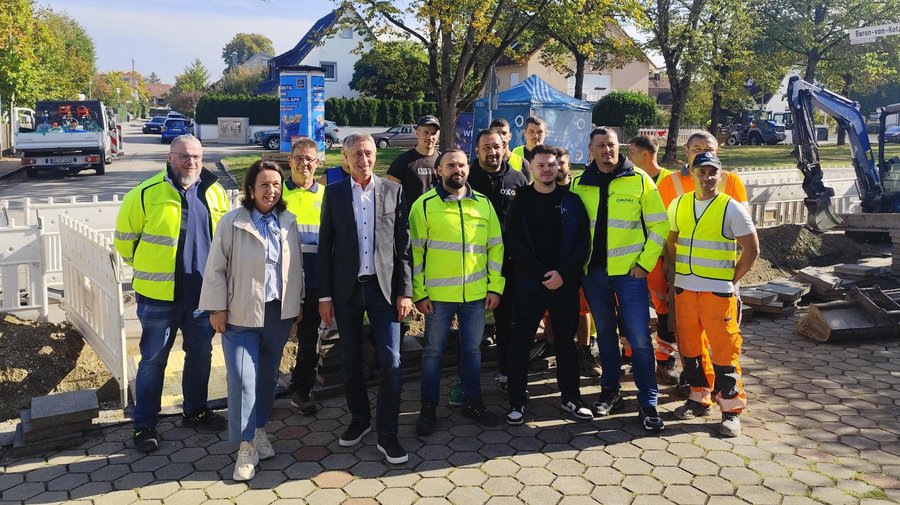 Gruppenfoto an einer Straßenbaustelle: Mehrere Personen stehen vor einer abgesperrten Baugrube mit Bagger und Baumaterialien. Einige tragen Warnwesten. Im Hintergrund sind Bäume, Wohnhäuser und ein Informationsstand zu sehen.