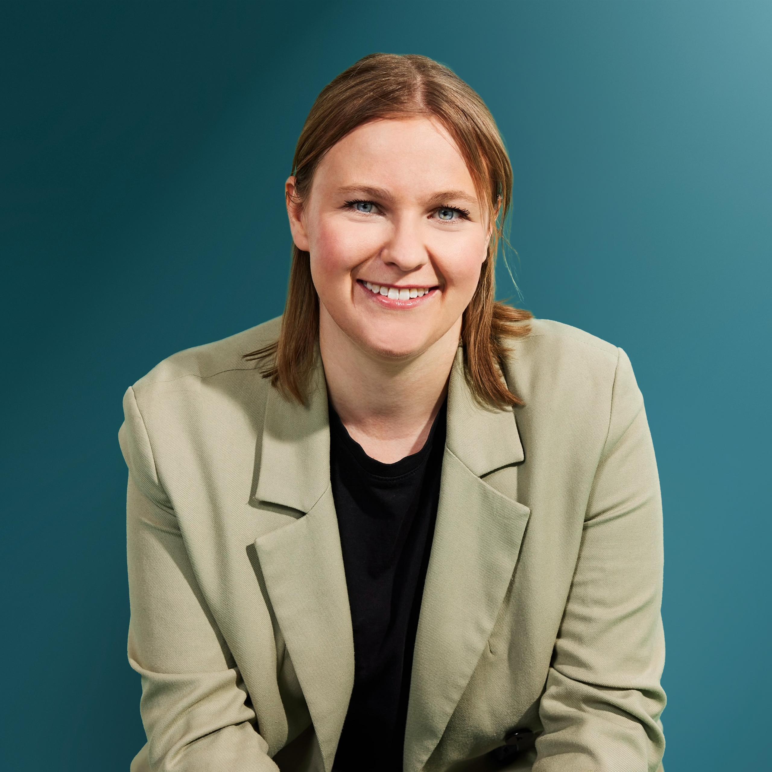 Lächelnde Frau mit kurzen, blonden Haaren in einem hellen Blazer vor einem türkisfarbenen Hintergrund