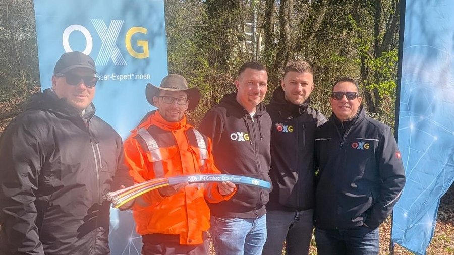 Fünf Personen stehen draußen für ein Gruppenfoto, teils in Bauarbeitskleidung, teils in OXG‑Kleidung, vor OXG‑Bannern. Sie halten gemeinsam ein petrolfarbenes Glasfaserkabel.