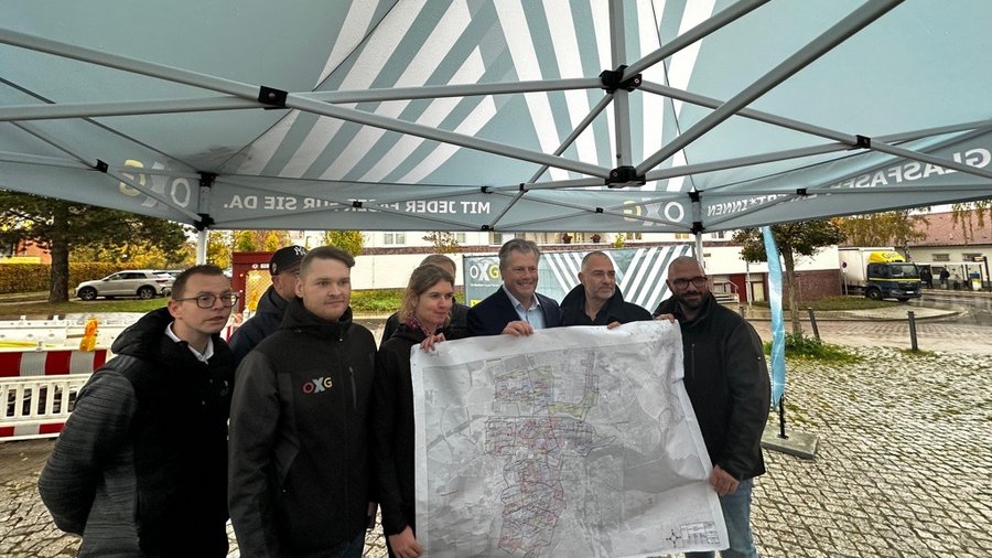 Eine Gruppe von fünf Personen steht an einer Baustelle in Weimar unter einem Zelt und hält eine große Karte, während im Hintergrund eine belebte Straße zu sehen ist.
