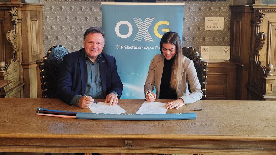 Zwei Personen sitzen an einem Tisch in Kaufbeuren und unterzeichnen Verträge, während im Hintergrund ein Banner mit der Aufschrift "OXG Die Glasfaser-Expert*in" zu sehen ist.