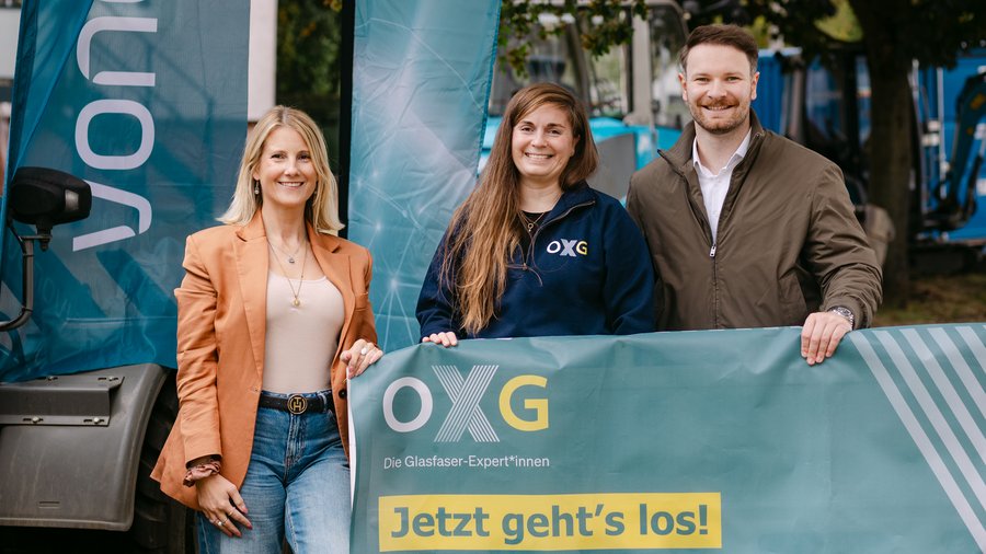 Drei lächelnde Personen halten draußen ein Werbebanner mit der Aufschrift "Jetzt geht's los" hoch. Im Hintergrund sieht man Werbemittel von Vonovia und OXG. Anlass ist der Baustart des Glasfaserprojekts in Hannover. 