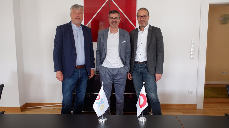 Drei Männer in formeller Kleidung stehen an einem Tisch mit Flaggen in einem modernen Konferenzraum