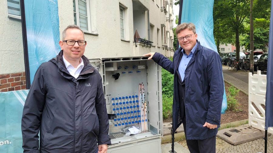 Zwei Männer stehen neben einem offenen Verteilerkasten im Freien in Treptow-Köpenick, während im Hintergrund ein Banner und einige Fahrräder zu sehen sind