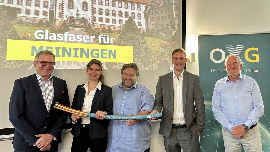 Eine Gruppe von fünf Personen steht in einem Raum vor einem Banner mit dem Schriftzug "Die Glasfaser-Expert*innen" und einer Präsentation, die sich auf "Glasfaser für MEININGEN" konzentriert, während sie Kabel halten.
