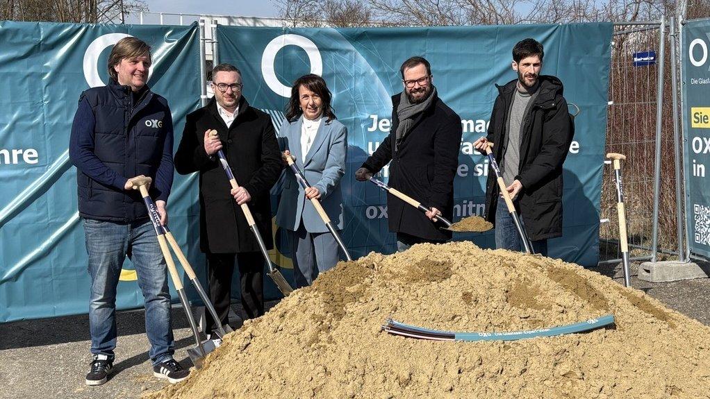 Fünf Personen stehen mit Spaten vor einem Sandhaufen bei einem symbolischen Baustart, im Hintergrund ein OXG‑Banner.
