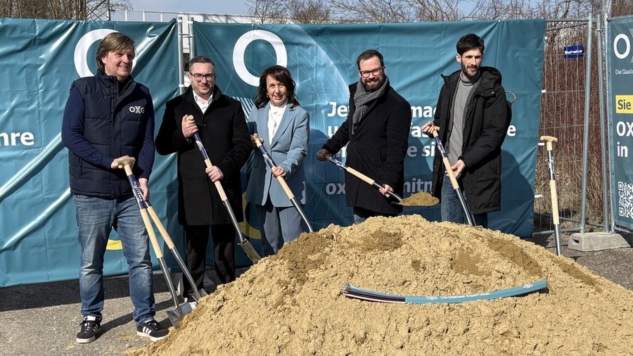 Fünf Personen stehen mit Spaten vor einem Sandhaufen bei einem symbolischen Baustart, im Hintergrund ein OXG‑Banner.