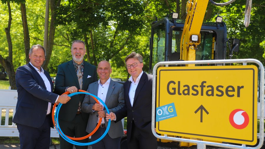 Vier Männer stehen lächelnd vor einem Bagger in Erfurt und halten orangefarbene und blaue Rohre in der Hand, neben ihnen befindet sich ein Schild mit der Aufschrift "Glasfaser"