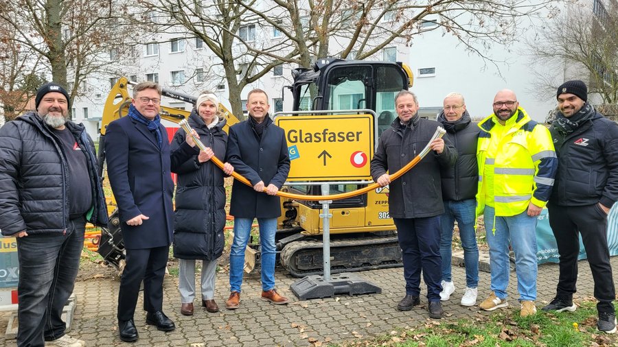Gruppenfoto von mehreren Personen, die eine Glasfaserleitung in der Hand halten, vor einem Baustellenschild und einem Bagger, während sie an einem Glasfaserprojekt
