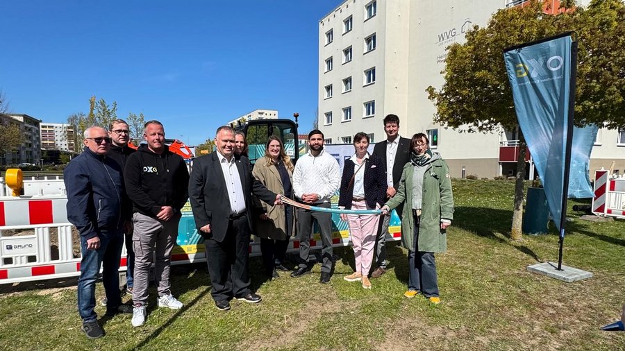 Eine Personengruppe draußen vor einer Glasfaserbaustelle. Einige Personen in der Mitte halten gemeinsam ein Glasfaserkabel. Im Hintergrund ein Wohngebäude.