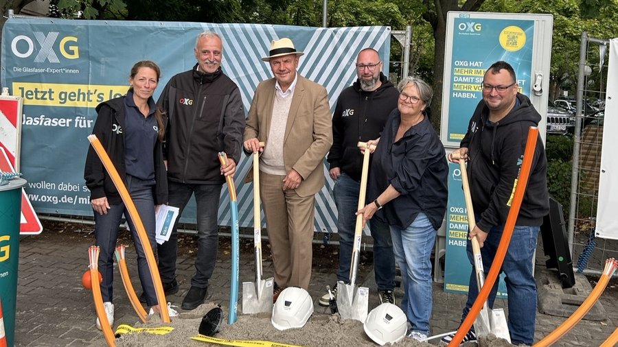 Gruppenfoto von sechs Personen, die Schaufeln halten und auf eine Baustelle in der Nähe eines Werbebanners der Firma OXG für Glasfaserangebote stehen