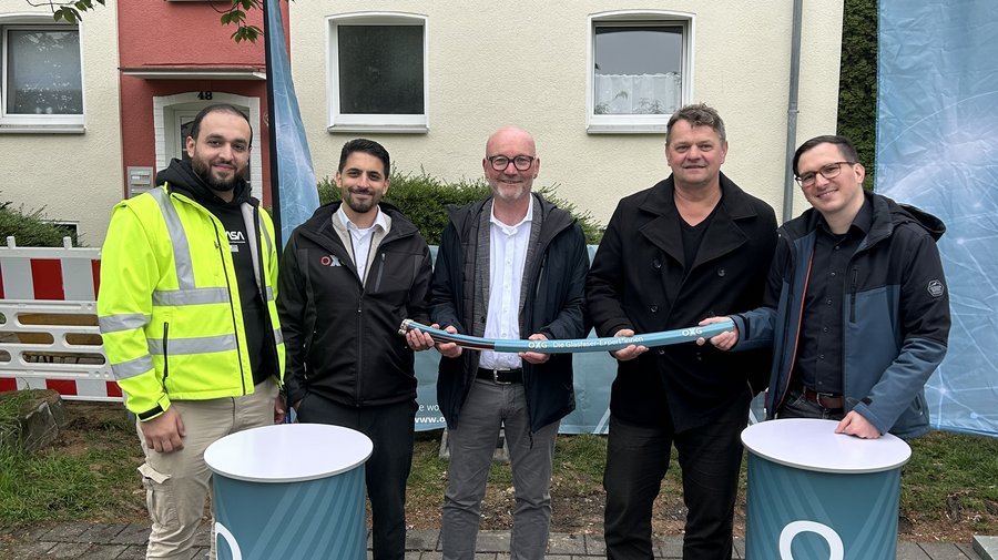 Eine Gruppe von fünf Männern nebeneinander zum gemeinsamen Foto. Sie stehen draußen vor einer Baustelle nahe eines Hauseingangs und halten ein Glasfaserkabel in die Kamera.