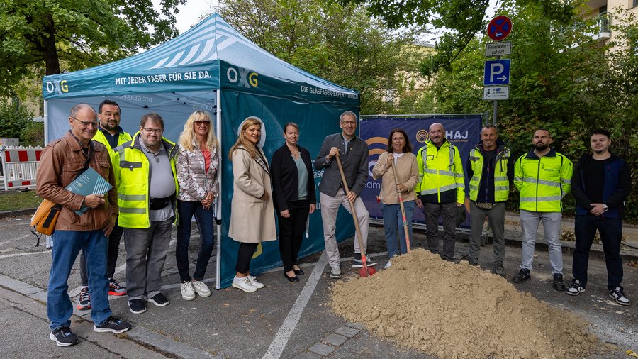 Vor einem OXG-Informationszelt stehen mehrere Personen neben einem Erdhaufen. Einige tragen Warnschutzkleidung, zwei halten Spaten. Die Aufnahme zeigt einen symbolischen Spatenstich zum Start eines Bauprojekts im Freien.