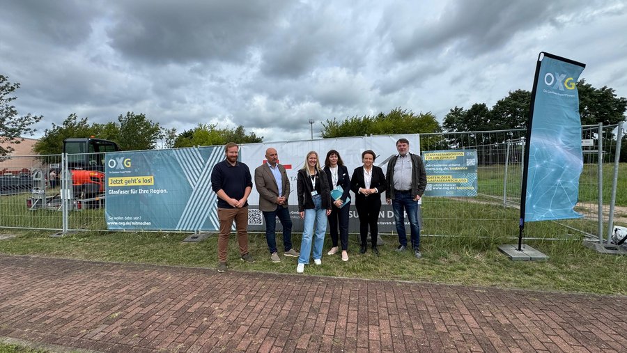 Eine Gruppe von sechs Personen steht vor einem Baustellenzaun mit Werbebannern für den Glasfaseranschluss in der Region Neustadt-Glewe, im Hintergrund sind Bäume und ein Baufahrzeug sichtbar