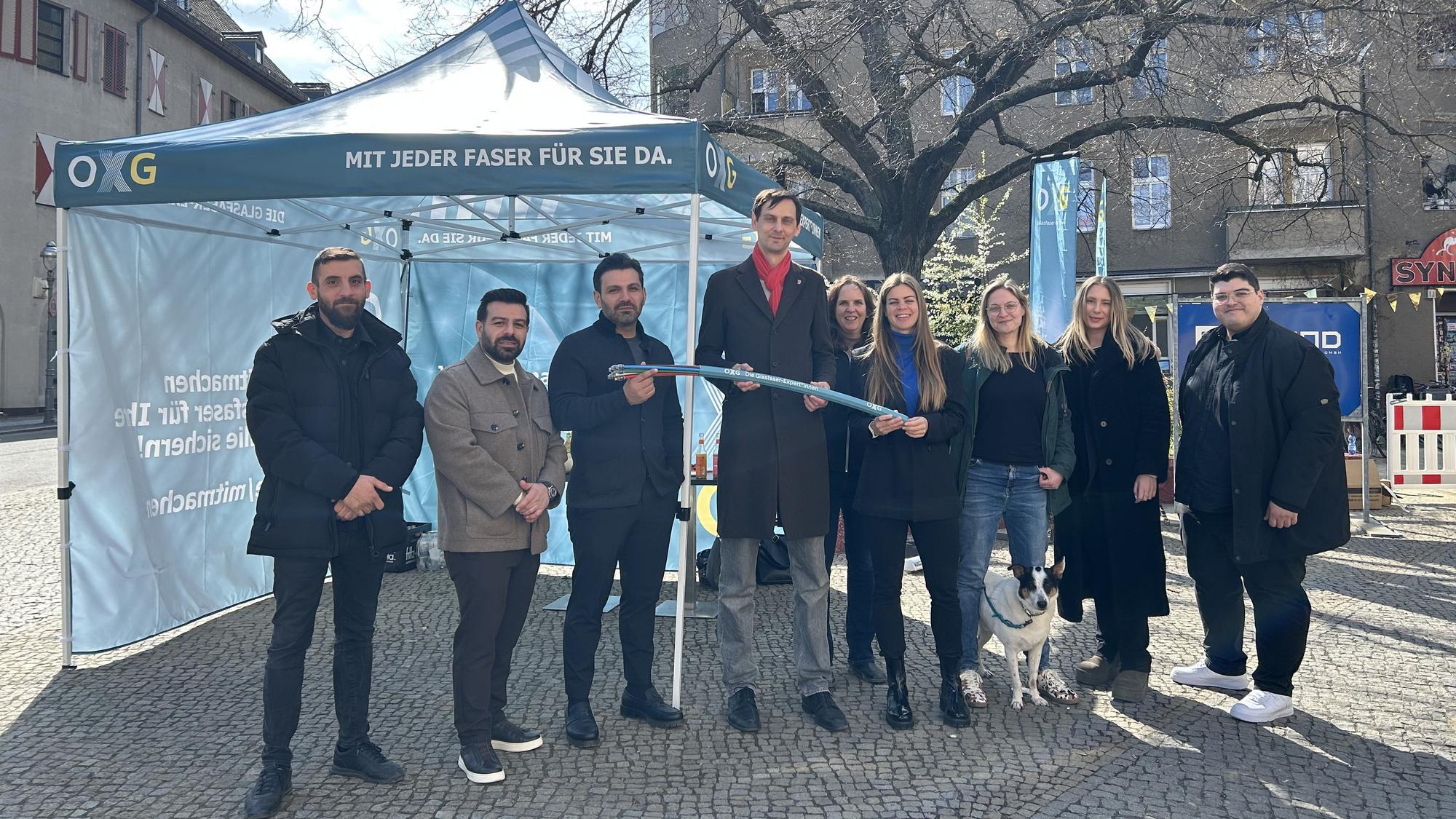 Eine Personengruppe steht draußen vor einem petrolfarbenen OXG-Pavillion. Mehrere Personen halten ein Glasfaserkabel fest.