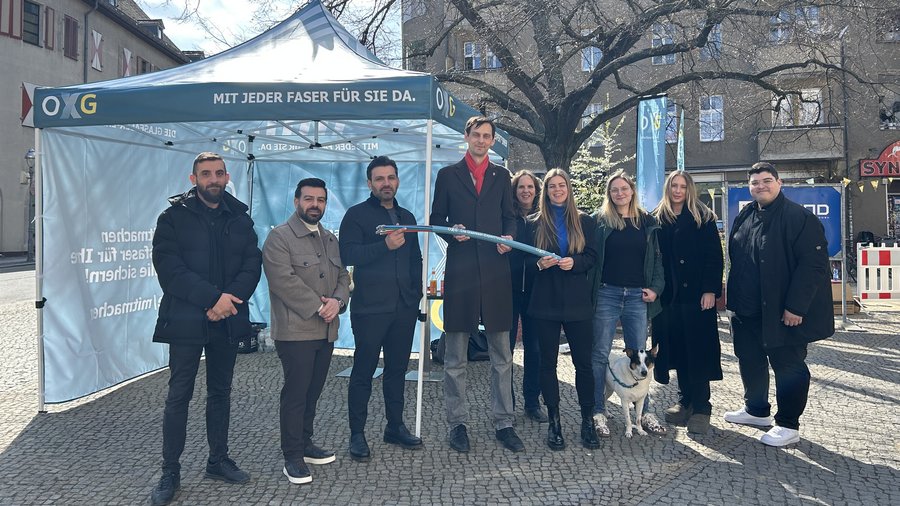 Eine Personengruppe steht draußen vor einem petrolfarbenen OXG-Pavillion. Mehrere Personen halten ein Glasfaserkabel fest.