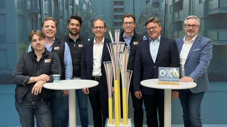 Gruppe von sechs Männern in Anzügen und Businessbekleidung, die vor einer modernen Architektur stehen und an zwei Stehtischen posieren, während sie neben einem Kunstobjekt aus bunten Rohren stehen