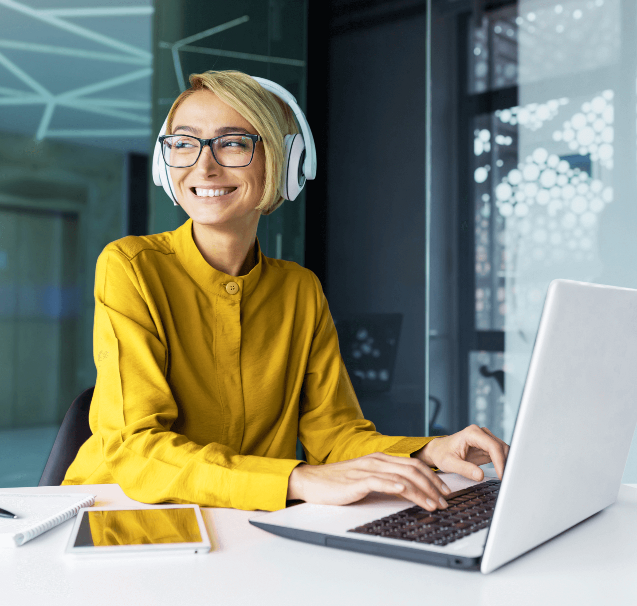 Lächelnde Frau mit blonden Haaren und Brille trägt Kopfhörer und arbeitet an einem Laptop in einem modernen Büro