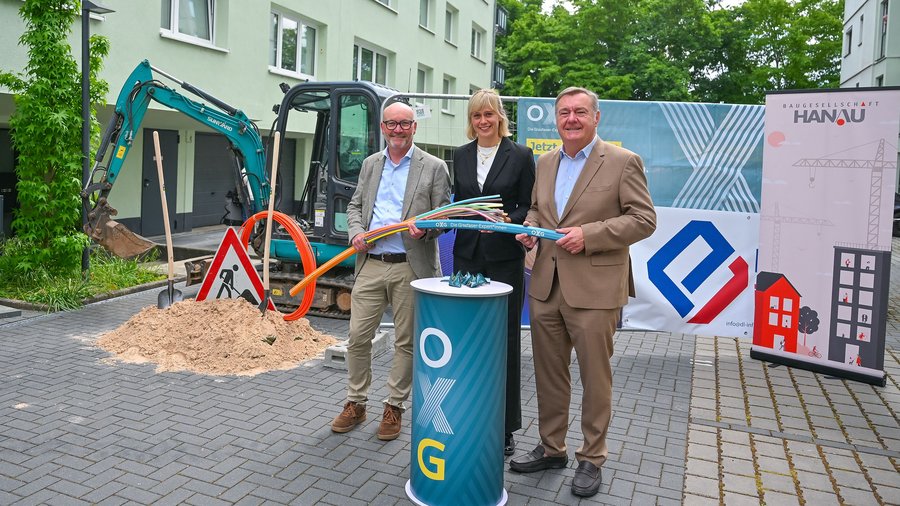 Drei Personen stehen vor einem Mehrfamilienhaus in Hanau neben einem Minibagger und einem aufgeschütteten Sandhaufen und halten bunte Glasfaser-Leerrohre in den Händen. Im Hintergrund sind Bauzäune mit den Logos von OXG und der Baugesellschaft Hanau sowie ein Baustellenschild zu sehen – symbolischer Start der Glasfaserbauarbeiten im Wohnquartier. Bildrechte: Stadt Hanau / Moritz Göbel