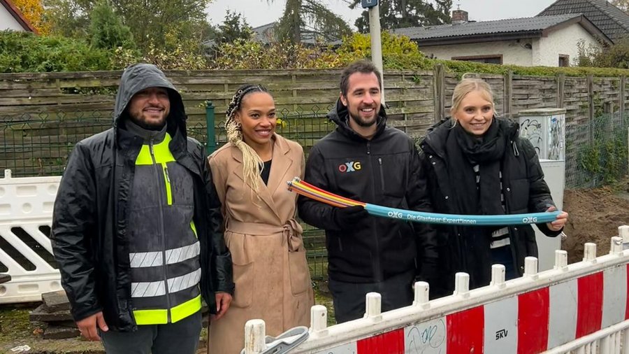 Vier Personen stehen lächelnd vor einer Baustelle in Lankwitz, während sie ein bunten Kabel in den Händen halten, umgeben von Absperrungen und einer ländlichen Kulisse.